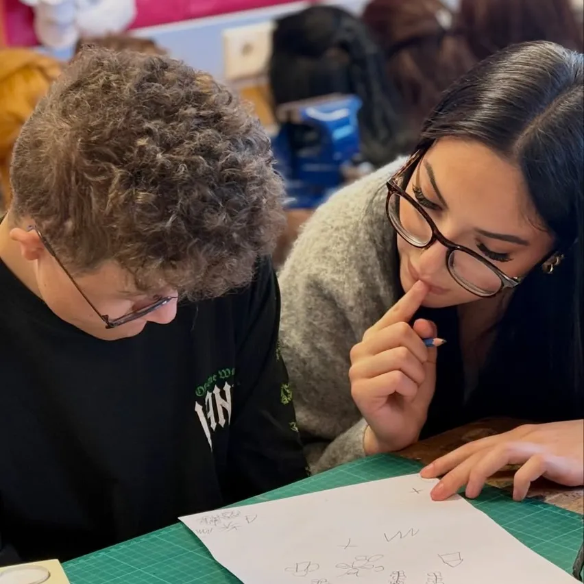 Zwei Schüler arbeiten zusammen an einer Zeichnung, einer mit lockigem Haar und einer Brille, der andere mit einer Brille und einem Pullover, beide konzentrieren sich auf das Papier vor ihnen.