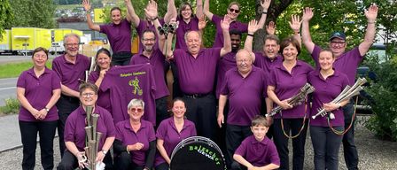 A group of musicians in purple shirts and black pants pose for a photo with their instruments in a park with a tree in the background.