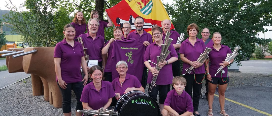 Eine Gruppe von Menschen in lila Shirts posiert für ein Foto mit Musikinstrumenten und einer Fahne. Die Fahne hat das Wort Balgacher. Sie lächeln und scheinen sich in einem Park zu befinden.