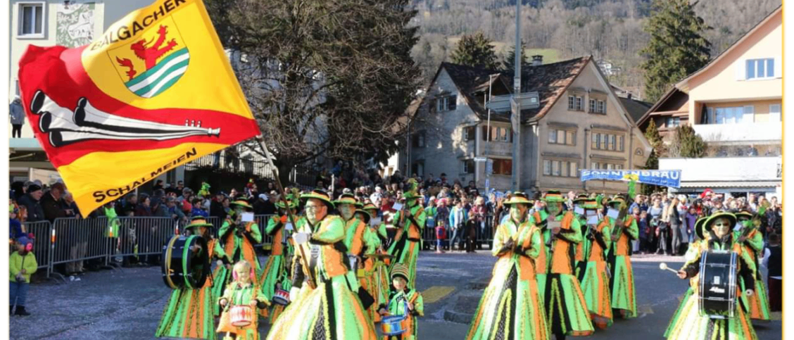 Ein Umzug von Menschen in grünen und orangefarbenen Kostümen, die Trommeln tragen. Eine Fahne mit Löwenemblem weht in der linken Ecke. Im Hintergrund sehen Zuschauer den Umzug.