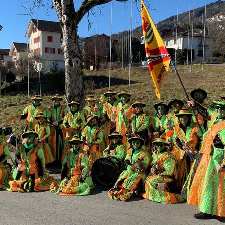 Eine Gruppe von Menschen in grünen und orangefarbenen Kostümen posiert für ein Foto mit Musikinstrumenten in einem Park. Ein Fahnenmast und ein Baum sind im Hintergrund zu sehen.