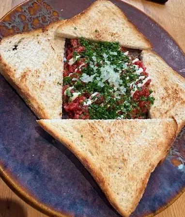 Ein quadratförmiges Gericht aus Fleisch, Käse und Kräutern zwischen Scheiben getoastetem Brot auf einem Teller.