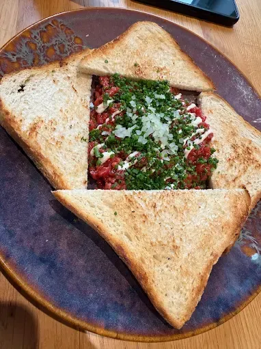 Ein quadratförmiges Gericht aus Fleisch, Käse und Kräutern zwischen Scheiben getoastetem Brot auf einem Teller.