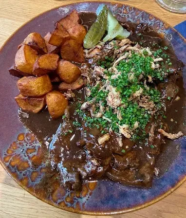 Ein Teller mit Rindergulasch mit gebratenen Kartoffeln, Frühlingszwiebeln und brauner Sauce steht auf einem Holztisch.