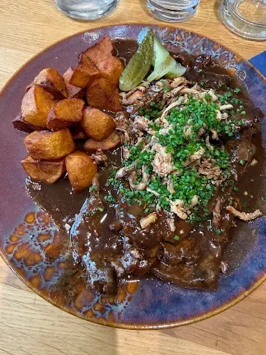 Ein Teller mit Rindergulasch mit gebratenen Kartoffeln, Frühlingszwiebeln und brauner Sauce steht auf einem Holztisch.