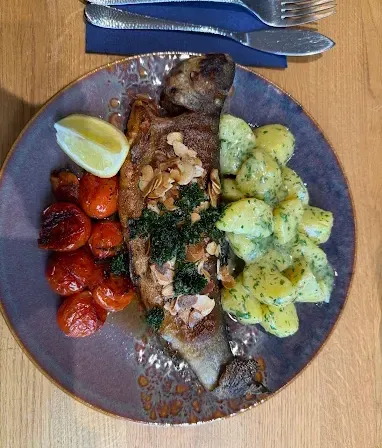 Ein ganzer Fisch auf einem Teller mit Kartoffelsalat, einer Zitronenscheibe und gerösteten Tomaten. Besteck auf einem blauen Serviettentuch.