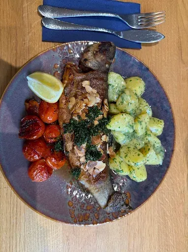 Ein ganzer Fisch auf einem Teller mit Kartoffelsalat, einer Zitronenscheibe und gerösteten Tomaten. Besteck auf einem blauen Serviettentuch.