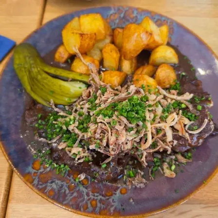 Ein Teller mit gebratenen Kartoffeln, einem eingelegtem Gurkenstück und Fleisch mit grünen Zwiebeln auf einem Holztisch.