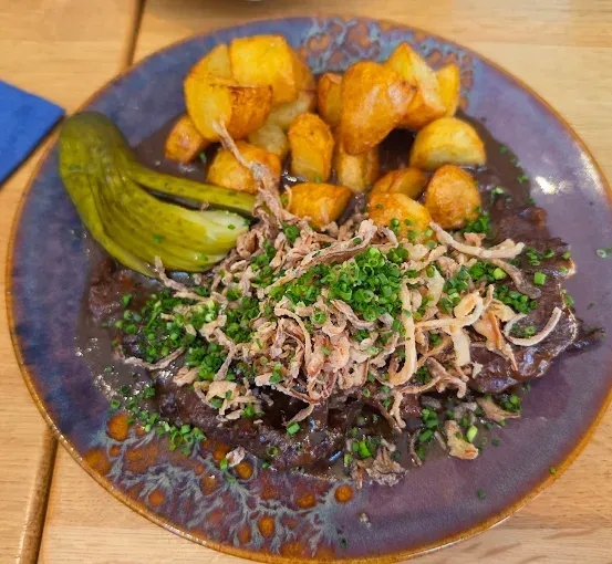 Ein Teller mit gebratenen Kartoffeln, einem eingelegtem Gurkenstück und Fleisch mit grünen Zwiebeln auf einem Holztisch.