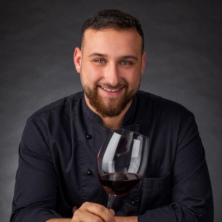 Ein Mann mit Bart in einer schwarzen Kochuniform hält ein Glas Rotwein und lächelt für die Kamera.