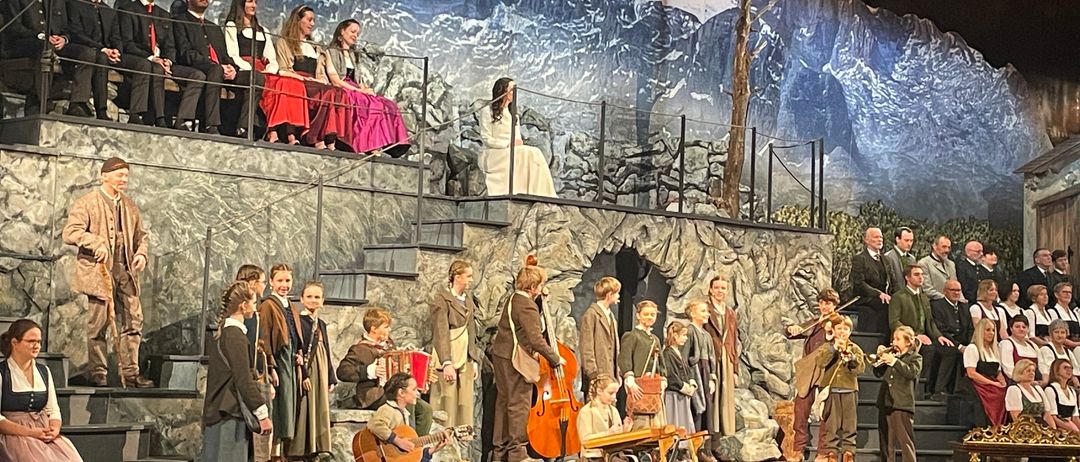 Eine Gruppe von Musikern, darunter Kinder, tritt auf der Bühne mit einer Kulisse von Berg und Burg bei Nacht auf. Einige sitzen, andere stehen mit Instrumenten wie Geigen und Cellos. Ein Publikum sitzt davor.