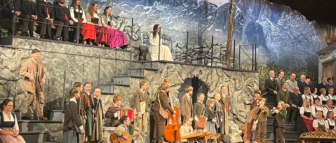 Eine Gruppe von Musikern, darunter Kinder, tritt auf der Bühne mit einer Kulisse von Berg und Burg bei Nacht auf. Einige sitzen, andere stehen mit Instrumenten wie Geigen und Cellos. Ein Publikum sitzt davor.