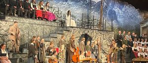 Eine Gruppe von Musikern, darunter Kinder, tritt auf der Bühne mit einer Kulisse von Berg und Burg bei Nacht auf. Einige sitzen, andere stehen mit Instrumenten wie Geigen und Cellos. Ein Publikum sitzt davor.