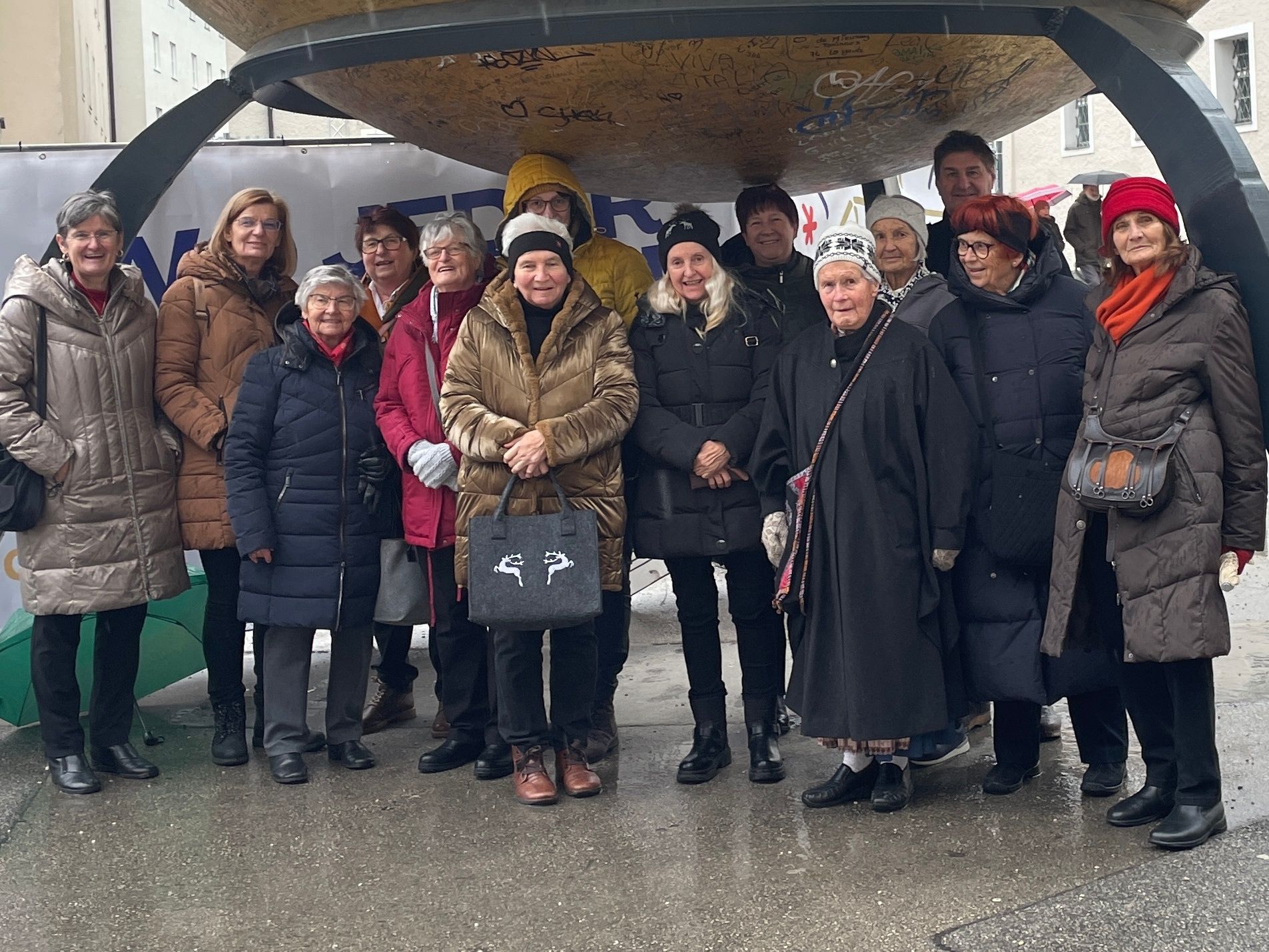 Eine Gruppe von Menschen steht vor einer großen, mit Graffiti bedeckten Struktur, unter einem Banner, bei regnerischem Wetter.