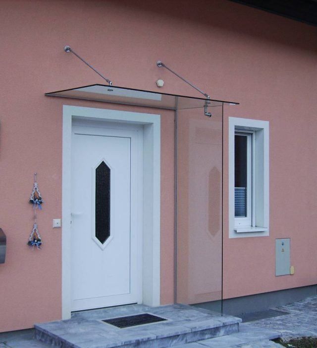 Ein modernes rosafarbenes Haus mit einer weißen Tür und einem Glasvordach darüber. Das Glasvordach hat Metallstützen.