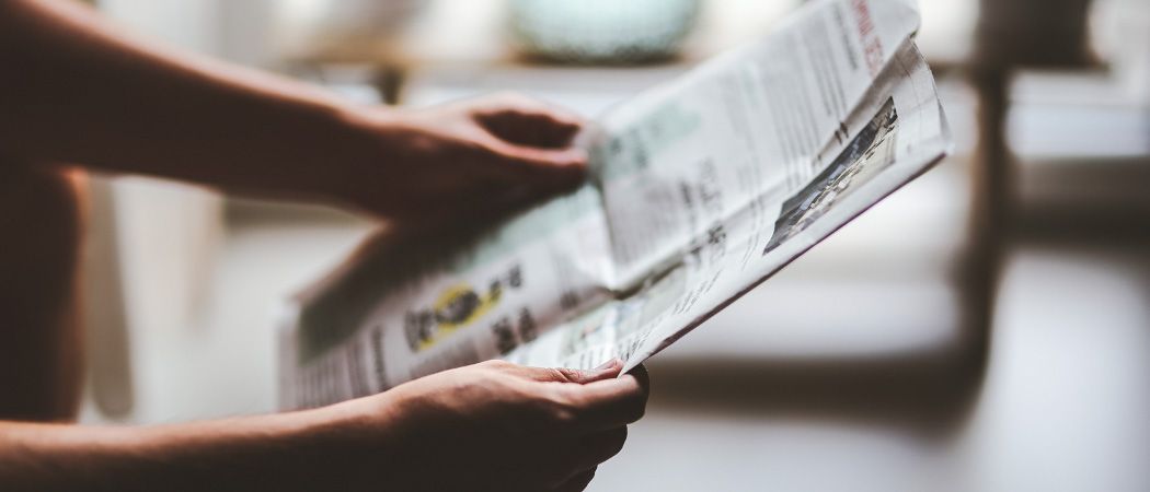Eine Person hält eine Zeitung mit einem Bild einer Person und Text im Hintergrund. Die Zeitung hat einen gelben Kreis mit Text und ist gefaltet. Die Person hält die Zeitung mit beiden Händen. Der Schatten der Person spiegelt sich auf der Zeitung. Der Hintergrund ist unscharf. Die Hand der Person ist unscharf.