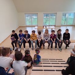 Gruppe von Kindern sitzt auf einer Bank in einem Raum mit großen Fenstern, einem Tisch und einem Holzboden.