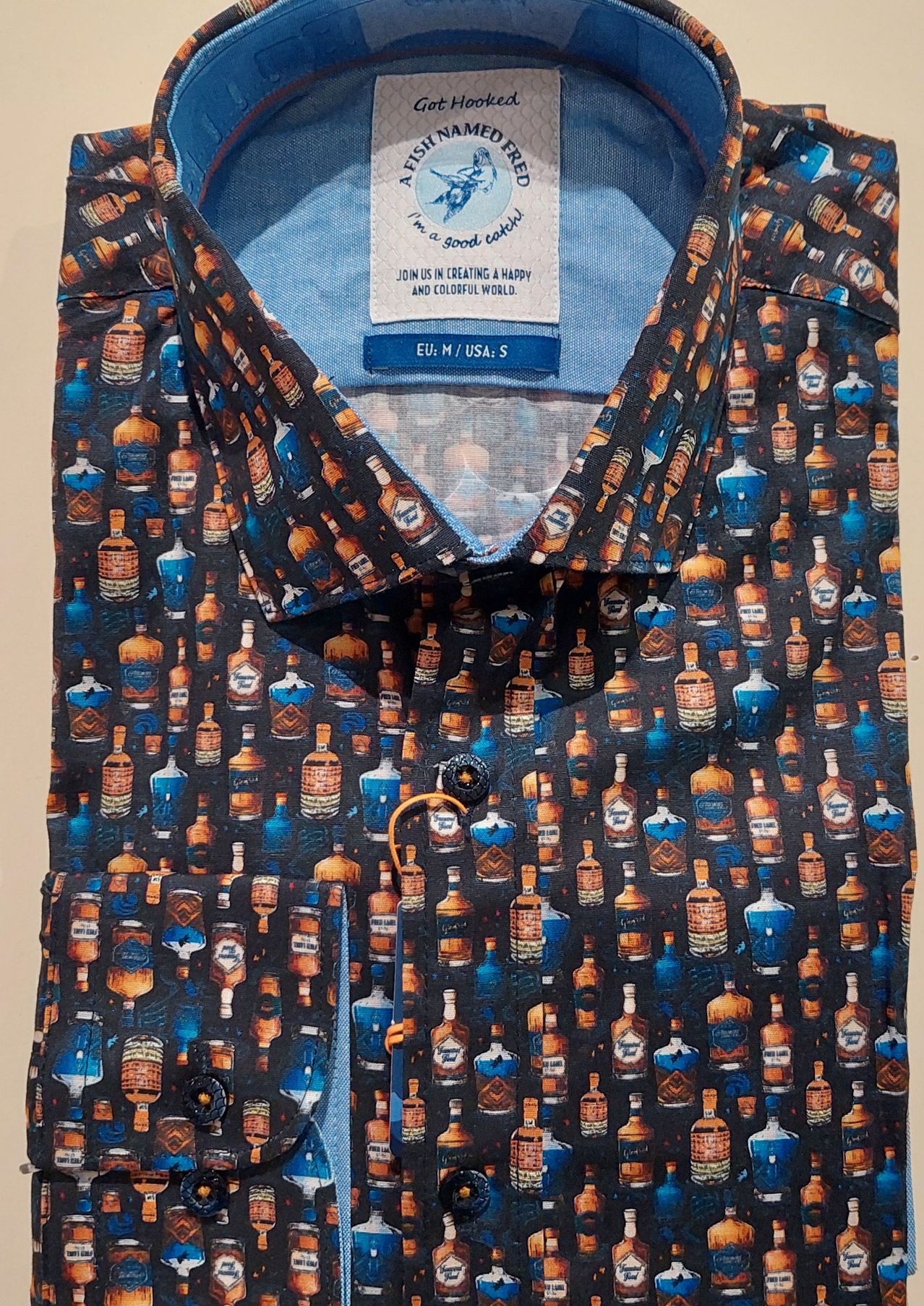 Ein Hemd mit einem Muster von Whiskyflaschen in Blau und Braun auf schwarzem Hintergrund. Der Kragen und die Knöpfe sind blau. Ein Etikett zeigt 'EU: M/USA: S' an.