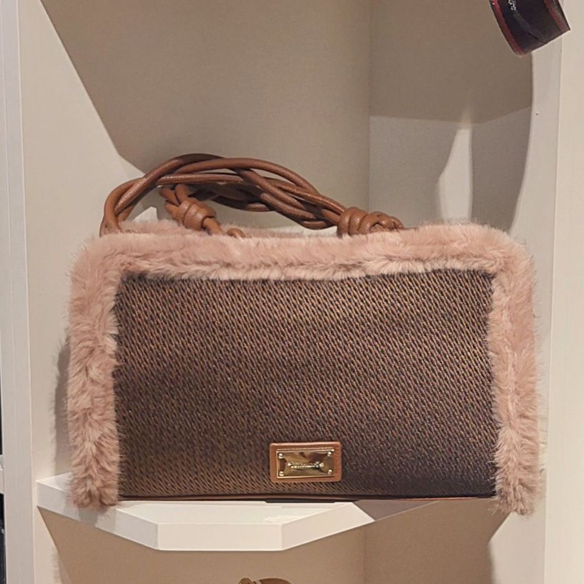 Eine braune Handtasche mit Pelzbesatz auf einem weißen Regal. Die Tasche hat eine gewebte Textur und ein goldfarbenes Logo.