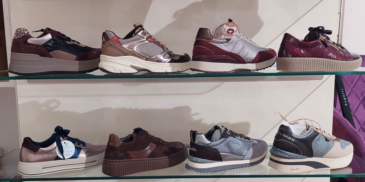 Vier Paar Sneaker auf einem Glasregal, darunter ein Paar in Marone und Silber, ein Paar in Braun und Weiß und ein Paar in Grau und Schwarz.