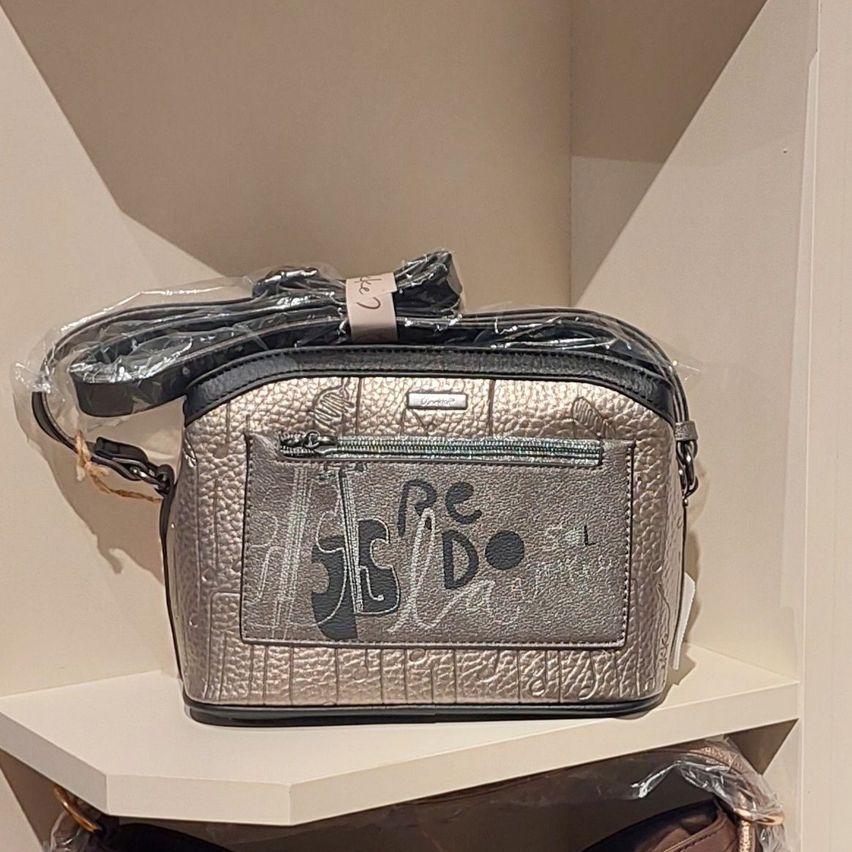A metallic silver handbag with a black strap and a logo on the front is displayed on a white shelf.