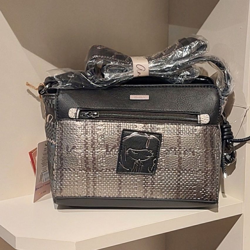 A black and silver handbag with a silver metallic pattern and a black leather patch with a dog design. The bag is wrapped in plastic and placed on a white shelf.