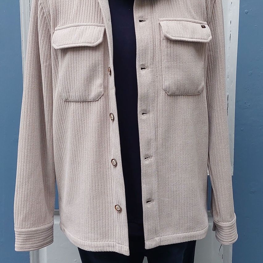 A light beige ribbed jacket with two chest pockets and buttoned front. The jacket is displayed on a mannequin against a blue background.