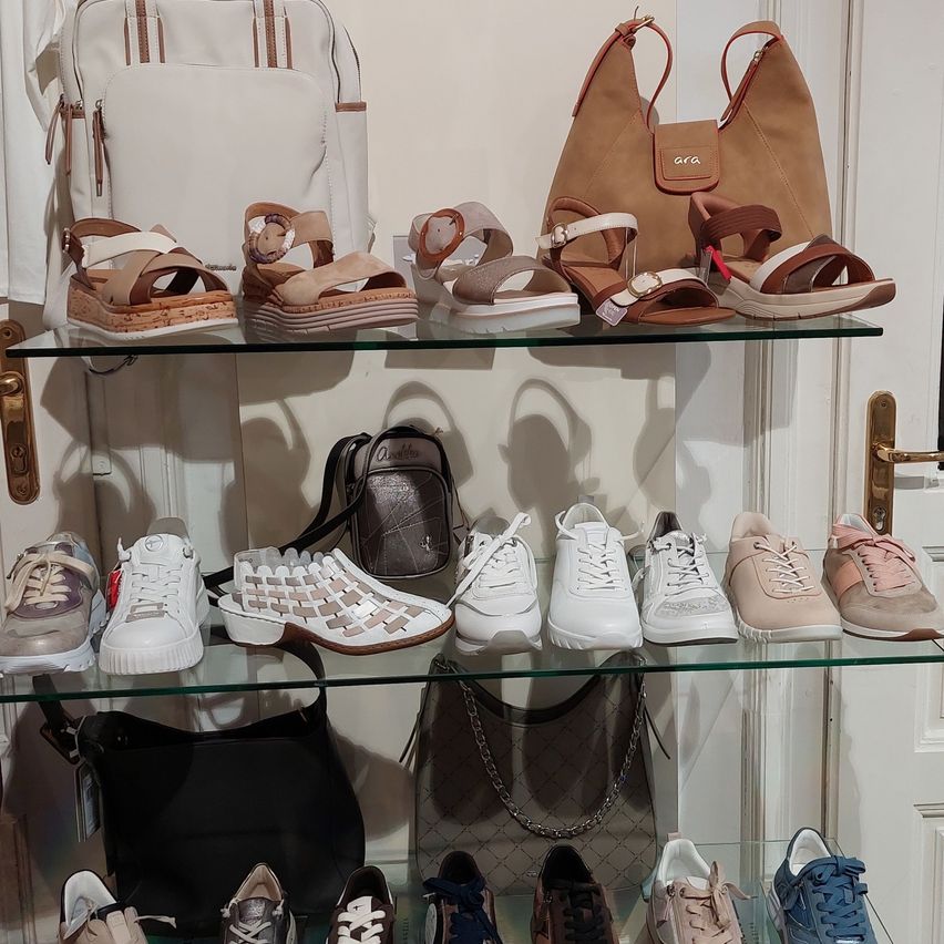 A glass shelf displays several pairs of sandals, sneakers, and bags. The sandals have straps, and some have wedge soles. The sneakers are in different colors, and some have laces. The bags are in different sizes and styles, and some have tags. The shelf is in front of a white door.