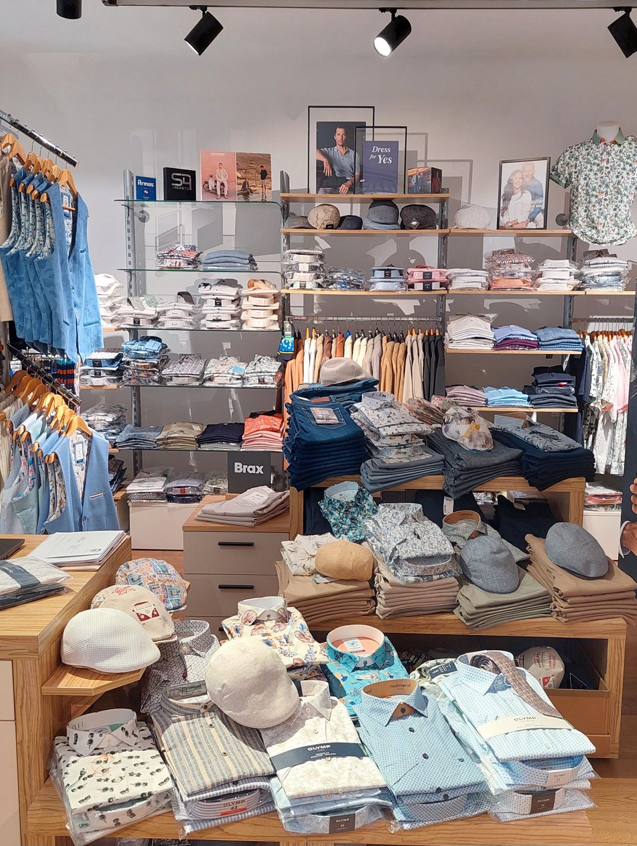 A clothing store with neatly arranged shelves displays various shirts, jeans, and hats. Mannequins and picture frames adorn the walls.