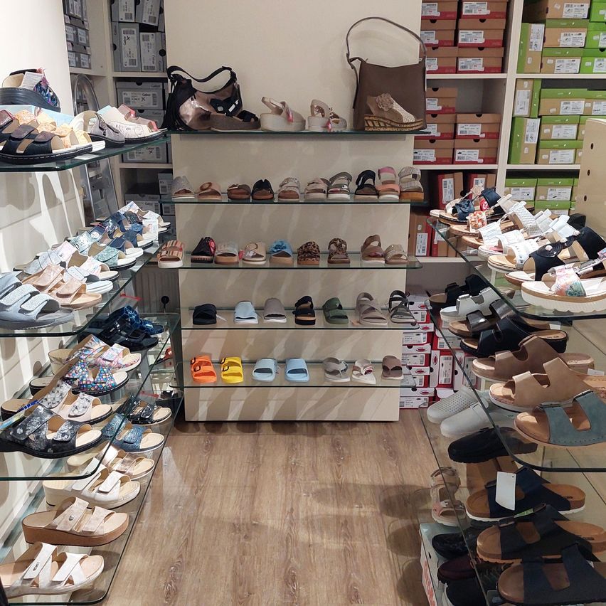 A shoe store filled with various sandals on glass shelves, wooden floor, and bags and boxes on the shelves.