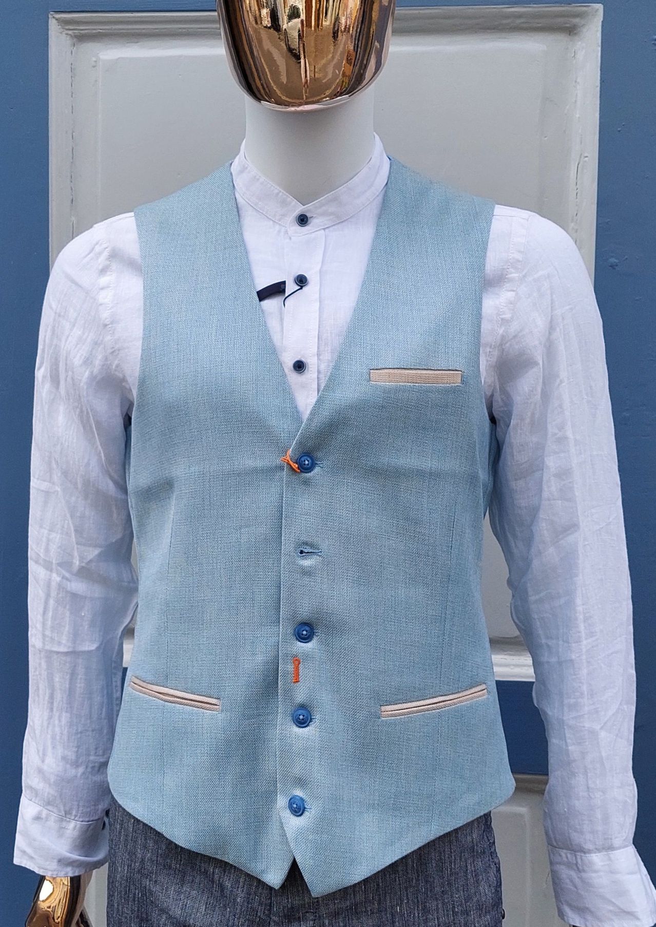 Ein Mannequin trägt eine hellblaue Leinenweste mit orangefarbenen Knöpfen und einer kleinen Tasche auf der linken Seite. Die Weste ist mit einem weißen Langarmhemd kombiniert.