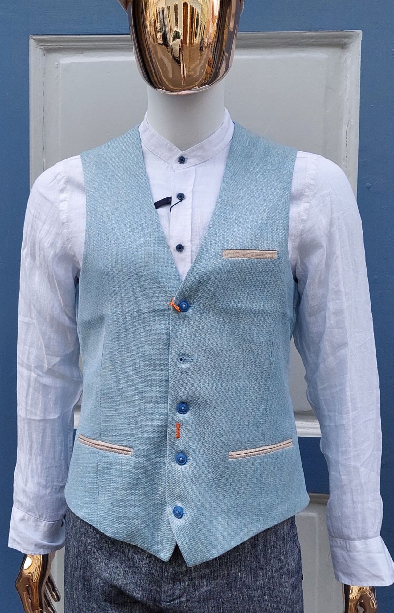 Ein Mannequin trägt eine hellblaue Leinenweste mit orangefarbenen Knöpfen und einer kleinen Tasche auf der linken Seite. Die Weste ist mit einem weißen Langarmhemd kombiniert.