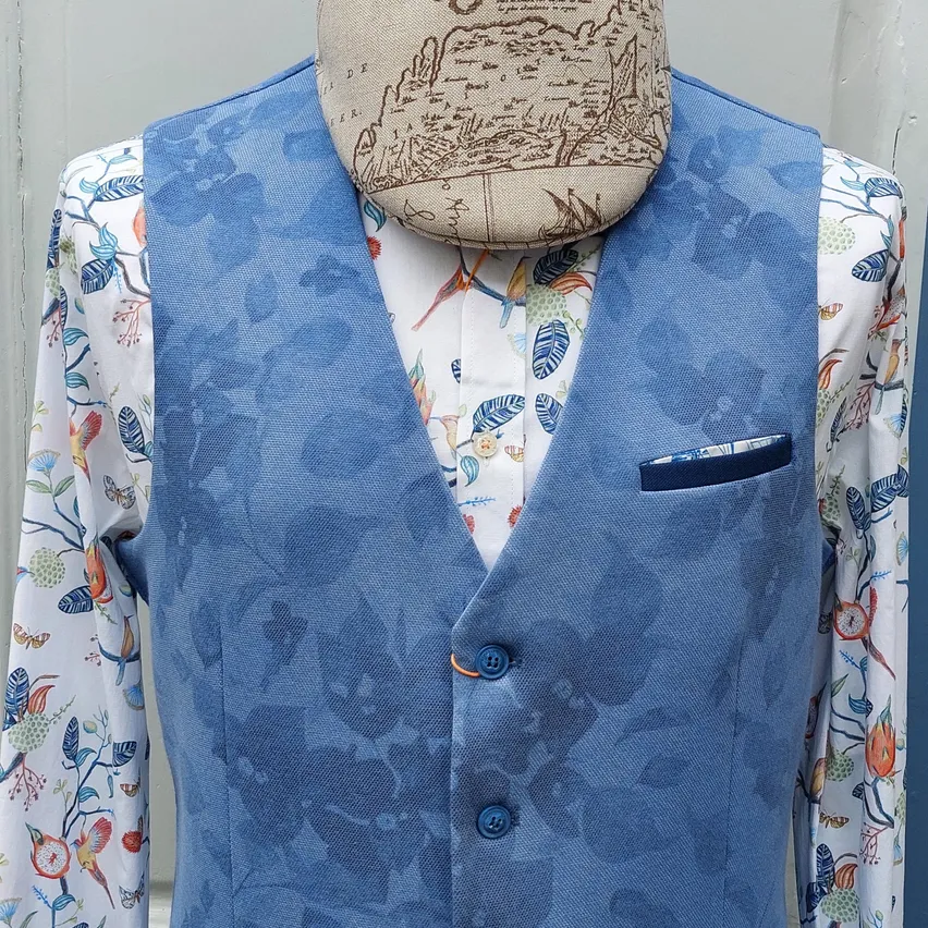 Ein Dummy zeigt eine blaue Weste mit Blumenmustern und einer Tasche auf einem blumig bedruckten Hemd mit einem beigefarbenen Hut auf dem Kopf.