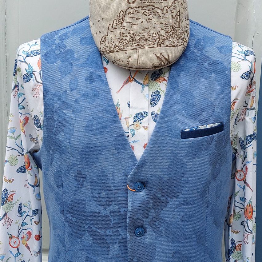 Ein Dummy zeigt eine blaue Weste mit Blumenmustern und einer Tasche auf einem blumig bedruckten Hemd mit einem beigefarbenen Hut auf dem Kopf.