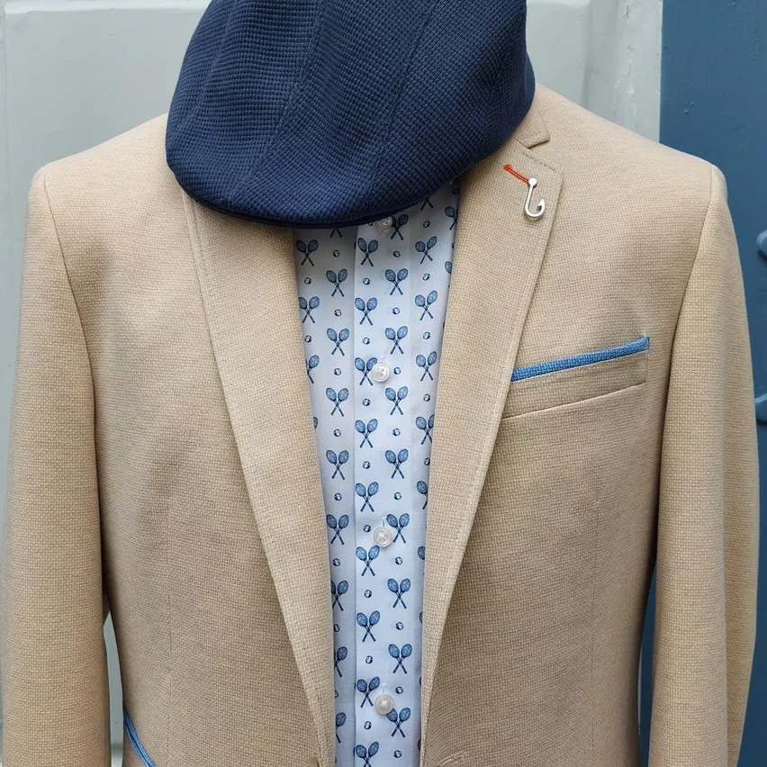 Ein beigefarbener Blazer mit blauem Innentaschenfutter und einer blauen Kappe steht auf einem Mannequin. Ein blauer Schal mit weißen Tennisschlägern ist darunter sichtbar.