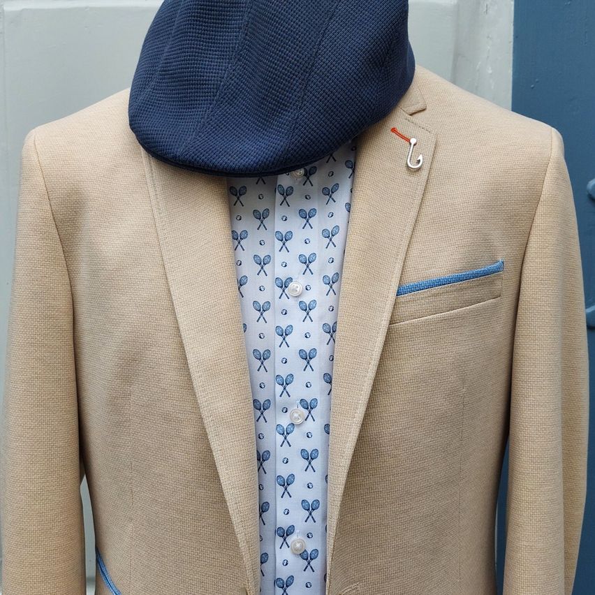 Ein beigefarbener Blazer mit blauem Innentaschenfutter und einer blauen Kappe steht auf einem Mannequin. Ein blauer Schal mit weißen Tennisschlägern ist darunter sichtbar.