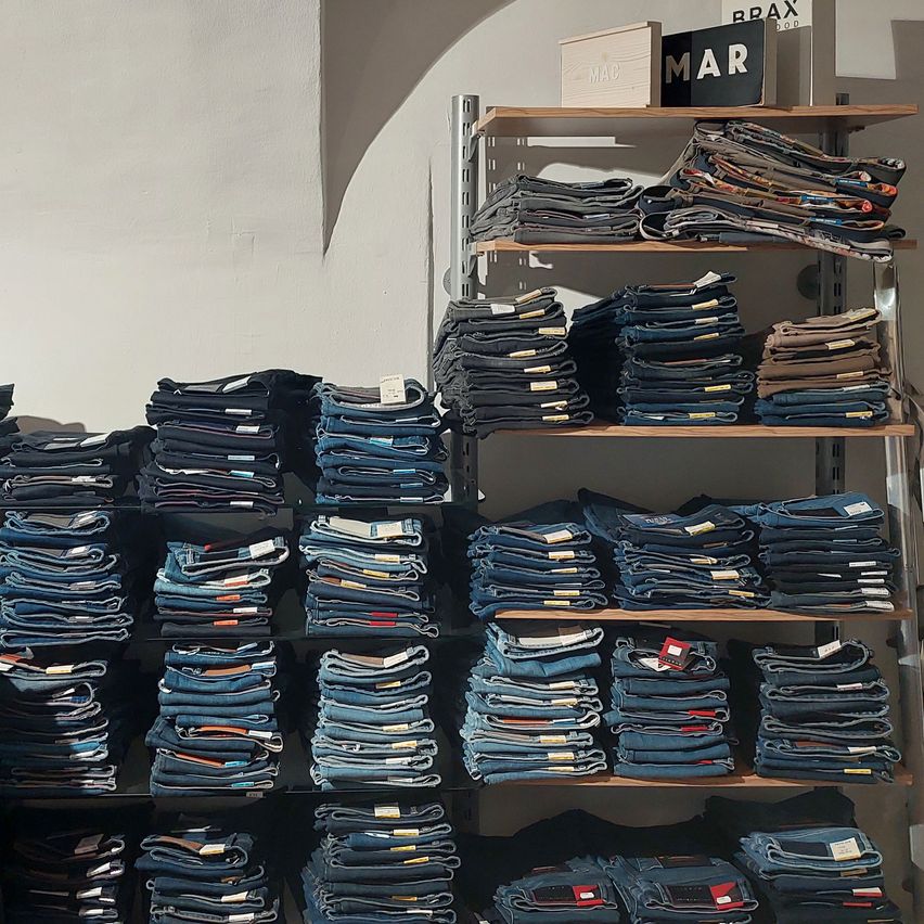 Ein Bekleidungsgeschäft zeigt mehrere Regale mit ordentlich gefalteten Jeans, mit einem Schild darüber, das MAR und ein anderes Schild, das BRAX liest.