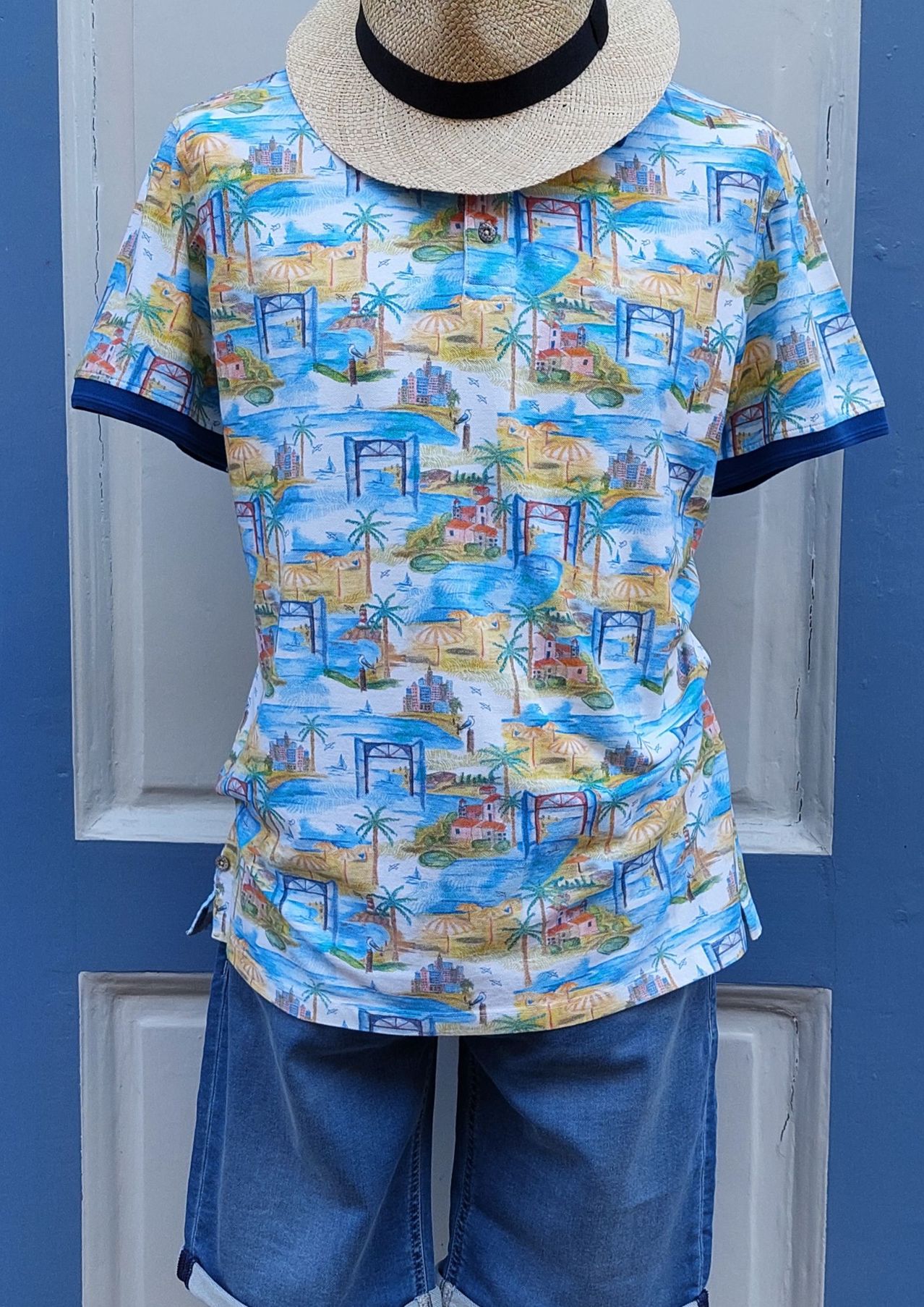 Ein Nahaufnahme eines blau-weiß bedruckten Poloshirts mit tropischem Landschaftsdesign, ausgestellt an einem Mannequin vor einer blauen Tür.