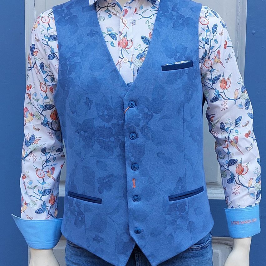 Ein Mannequin trägt eine blaue Weste mit Blumenmustern und ein weißes Langarmhemd mit passenden Blumendesigns. Die Weste hat blaue Knöpfe und Taschen.