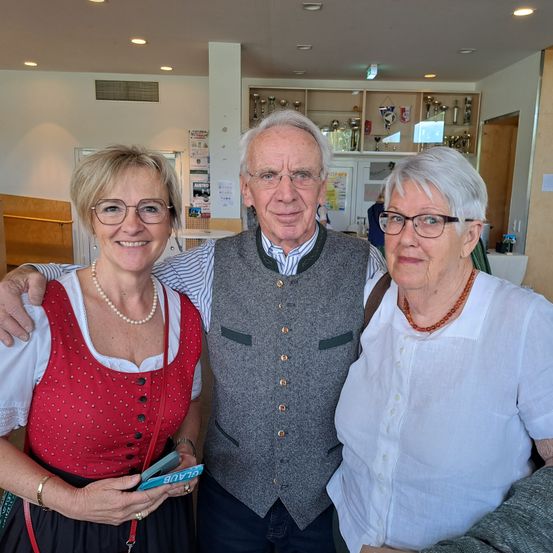 Drei Personen, ein Mann und zwei Frauen, stehen zusammen und lächeln für ein Foto. Der Mann ist in einem grauen Weste und gestreiftem Hemd gekleidet, während die beiden Frauen traditionelle Kleidung tragen. Die Frau auf der linken Seite hat ein rotes Kleid und eine Perlenkette, und die Frau auf der rechten Seite trägt eine weiße Bluse.
