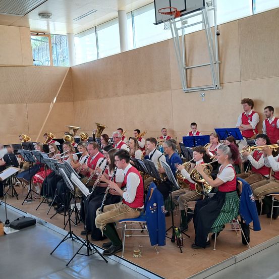 Eine Blaskapelle spielt auf einer Bühne. Mehrere Personen halten Instrumente, darunter Flöte, Trompete und Tuba. Sie sitzen auf Stühlen und stehen, einige tragen rote Westen. Vor ihnen stehen Notenständer.