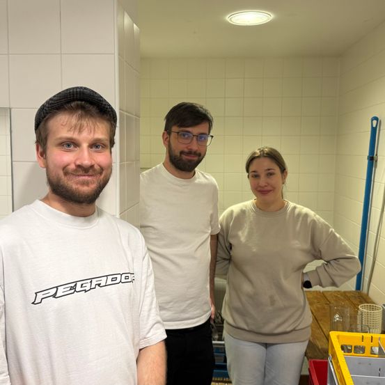 Drei Personen in einem kleinen Raum, lächelnd für ein Foto. Der Mann links trägt einen Hut und ein 'Pegado'-Shirt. Der Mann in der Mitte hat einen Bart und eine Brille. Die Frau rechts trägt einen beigen Pullover und weiße Hosen.