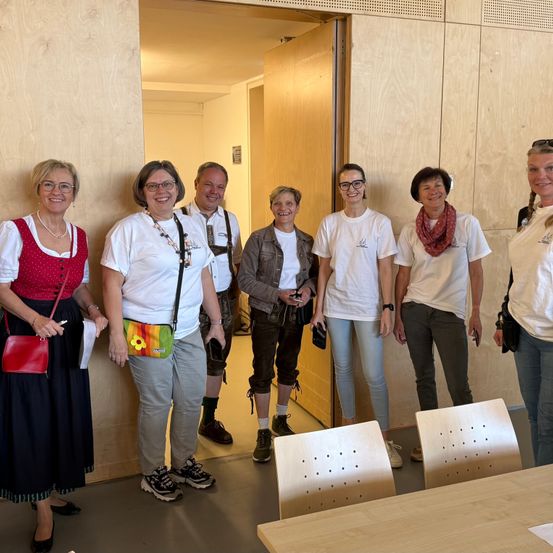Eine Gruppe Erwachsener steht in einem Raum mit Holzwänden. Sie tragen Freizeitkleidung, lächeln und posieren für ein Foto. Der Raum hat Stühle und einen Tisch. Die Frau in einem roten Kleid hält eine rote Tasche.