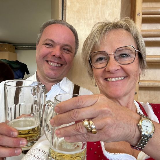 Ein Mann und eine Frau lächeln und halten Biergläser. Der Mann trägt ein weißes Hemd und Hosenträger, die Frau eine Brille und ein rotes Kleid. Sie sind in einem Raum mit Holzregalen und einer weißen Wand.