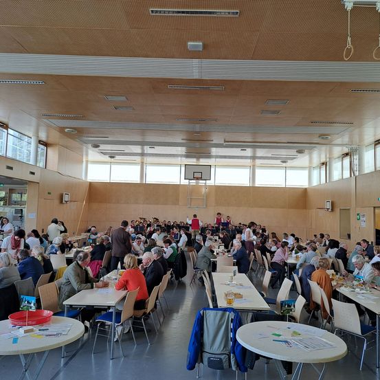 Ein großer Saal mit vielen Tischen, an denen Menschen sitzen und essen. Die Wände sind aus Holz, und es gibt Fenster mit großen Glasscheiben. An einer Wand befindet sich ein Basketballkorb.