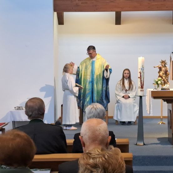 Ein Priester in einem farbenfrohen Gewand segnet zwei junge Mädchen. Sie stehen vor ihm und halten ein Buch und eine Kerze. Gläubige sitzen auf Bänken hinter ihnen und beobachten.