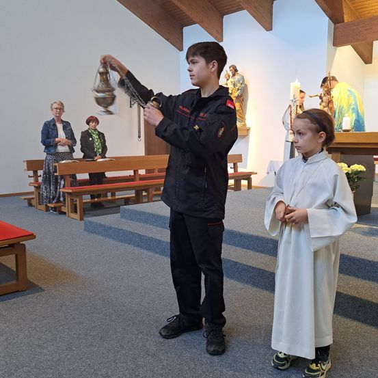 Ein Junge hält einen Räuchergefäß in einer Kirche, neben einem Mädchen in einer weißen Robe. Hinter ihnen sitzen Leute auf Bänken, und Statuen und Kerzen sind auf dem Altar.