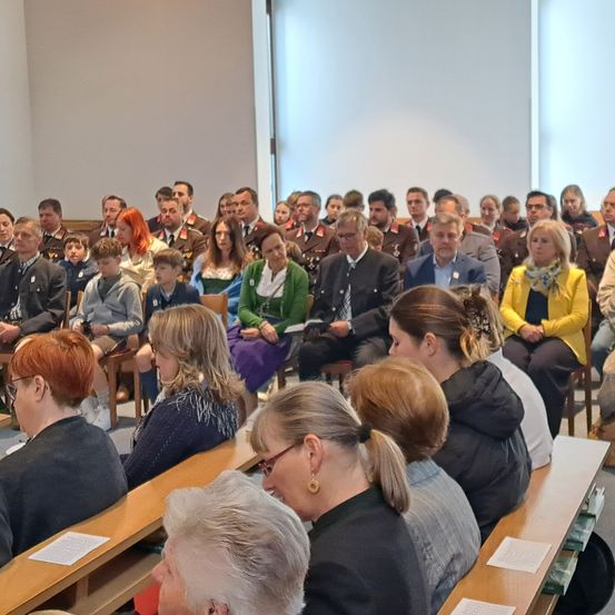 Eine Gruppe von Menschen versammelt sich in einem Raum für eine formelle Veranstaltung. Einige sitzen, andere stehen. Papiere liegen auf den Bänken, und einige tragen Uniformen.