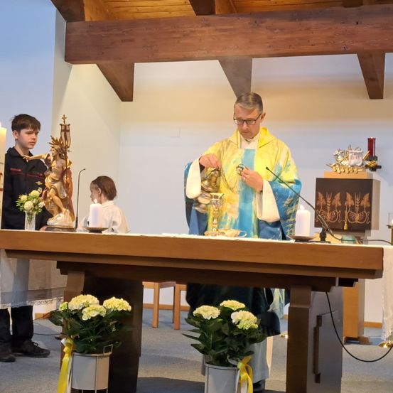 Ein Priester in einer blauen und goldenen Robe führt eine Zeremonie an einem Altar durch, mit einem Jungen und einem Mädchen an seiner Seite. Der Altar ist mit Kerzen und Blumen geschmückt.