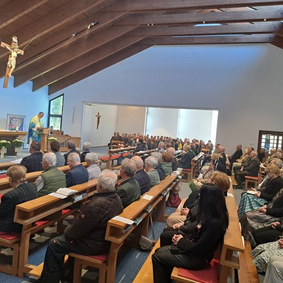 Ein Priester hält eine Rede in einer Kirche, in der viele Menschen auf Bänken sitzen. Ein Kreuz befindet sich an der Decke.